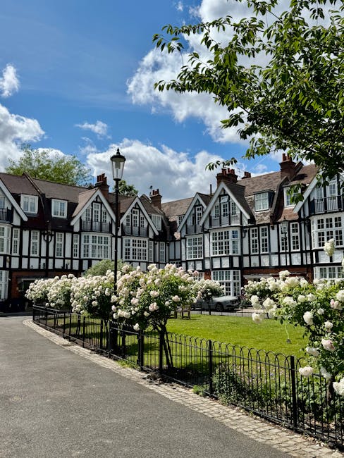 A row of picturesque terraced homes with traditional black and white timber-framed facades, set against a bright blue sky with scattered white clouds. In the foreground, a small, neatly maintained gar